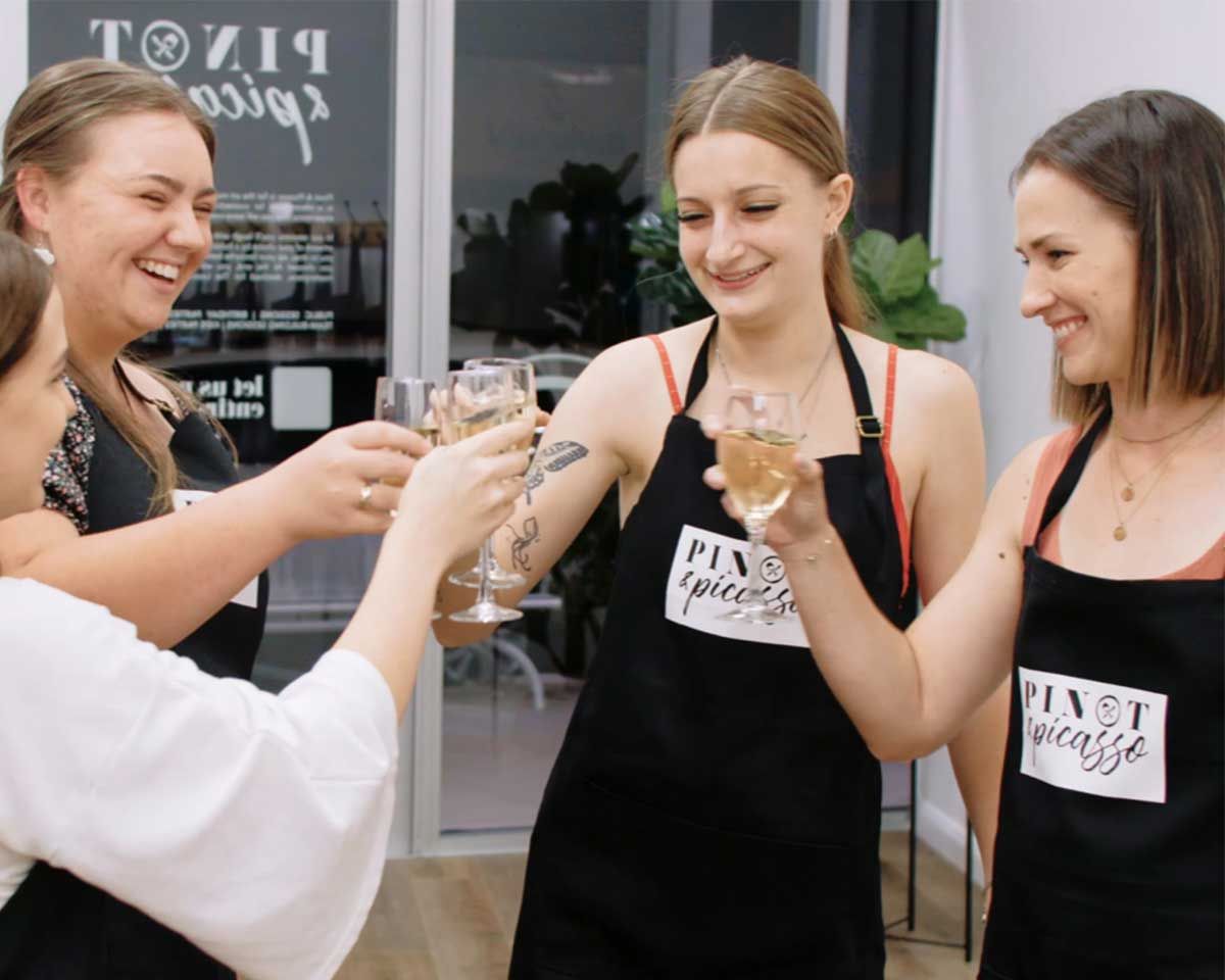 Four women celebrating at Pinot & Picasso with champagne saying cheers with their aprons on ready to paint and sip