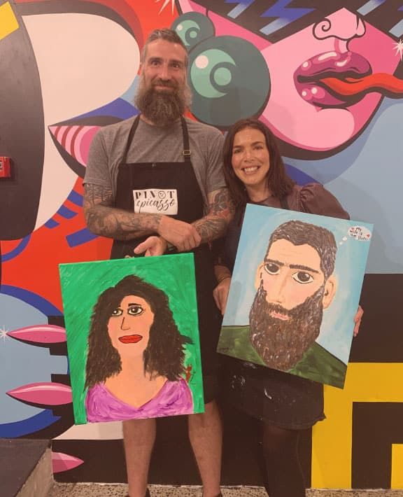 a man and a woman who are the franchisees of Pinot and Picasso Wellington stand with their aprons on and artworks they have painted of each other