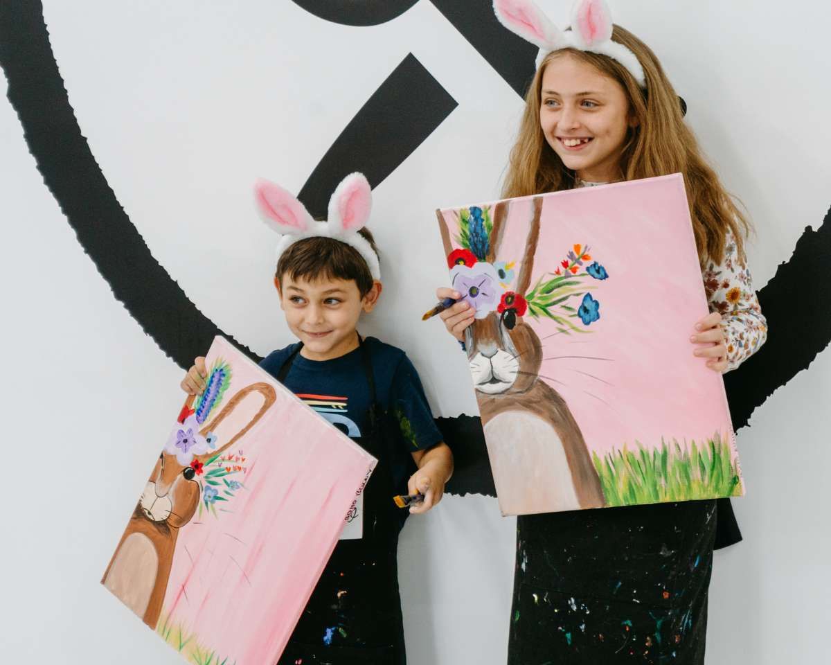a girl and a boy with bunny ears on are smiling holding the easter bunny artwork they painted at Pinot & Picasso