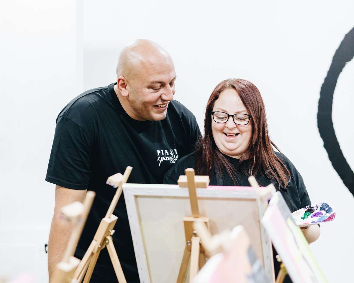 a man and a woman laughing and painting together at a Pinot and Picasso session