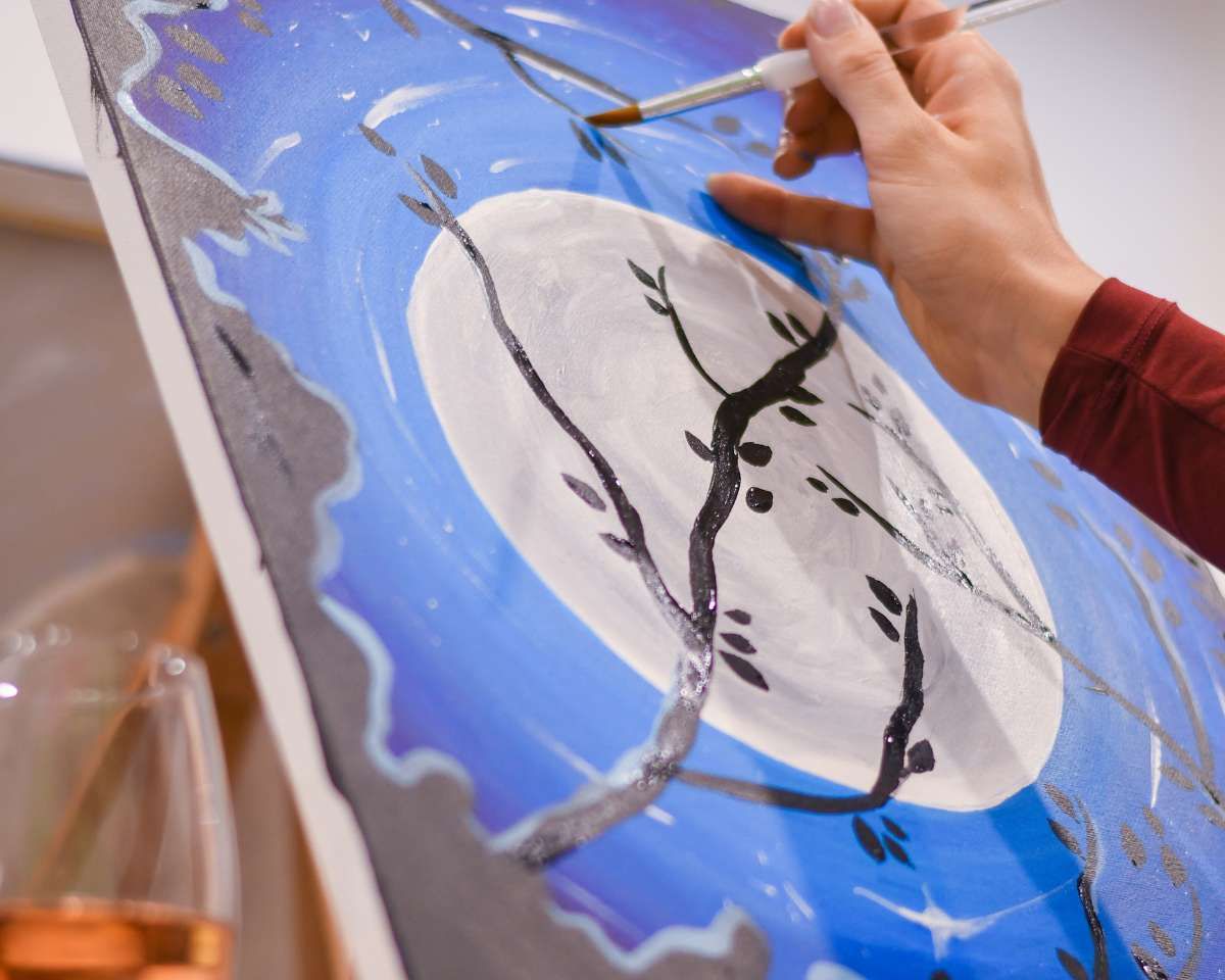 a person using a paintbrush to paint some tree branches in front of a moon at Pinot and Picasso