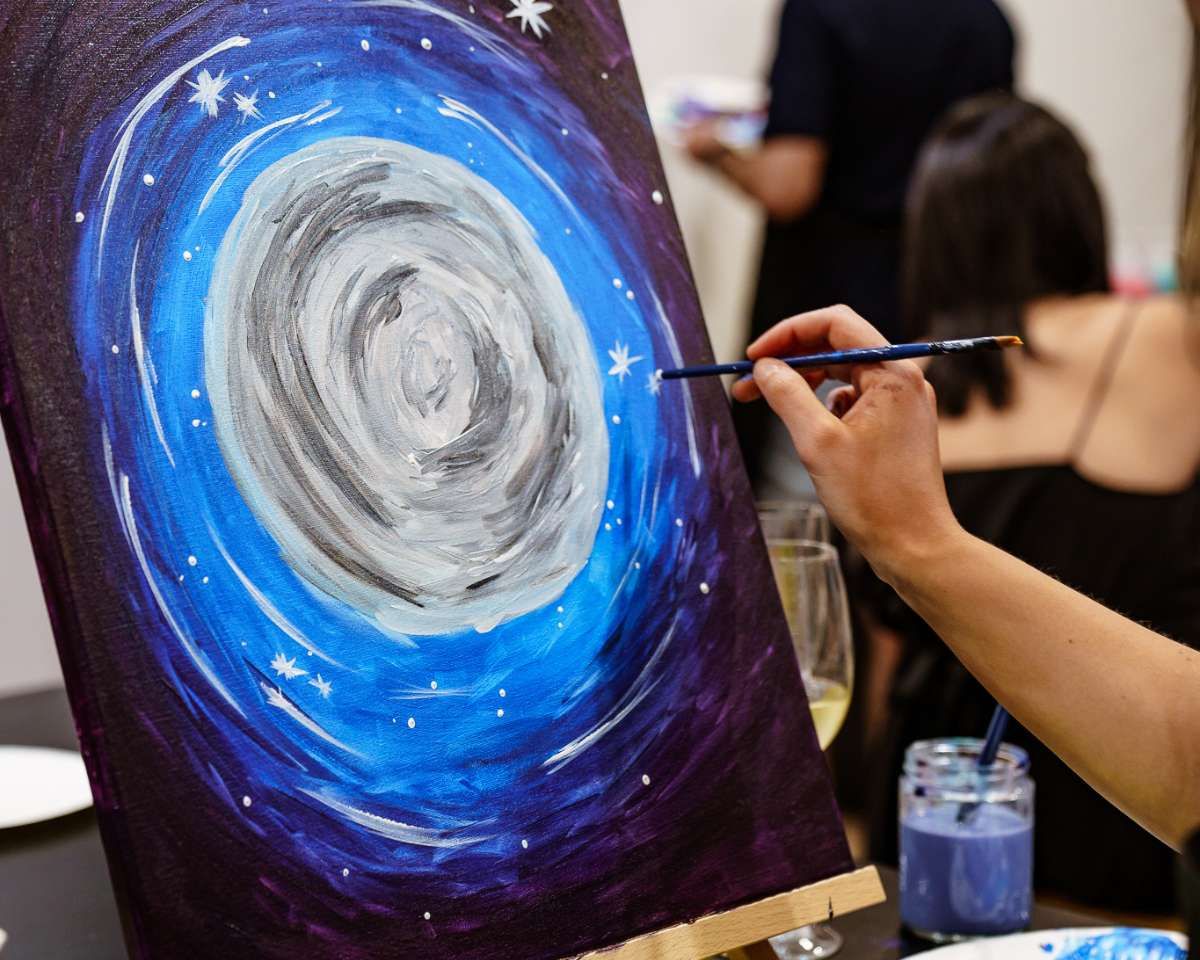 A woman attends a Pinot and Picasso paint and sip session to create a painting of a starry sky. A woman attends a Pinot and Picasso paint and sip session to create a painting of a starry night sky.