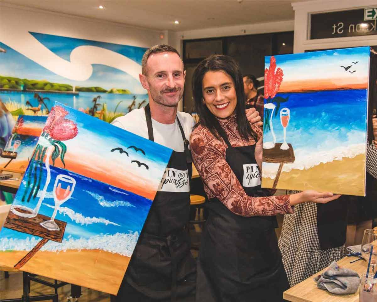 A man and woman posing for a photo with their artworks during a Pinot & Picasso paint and sip session.