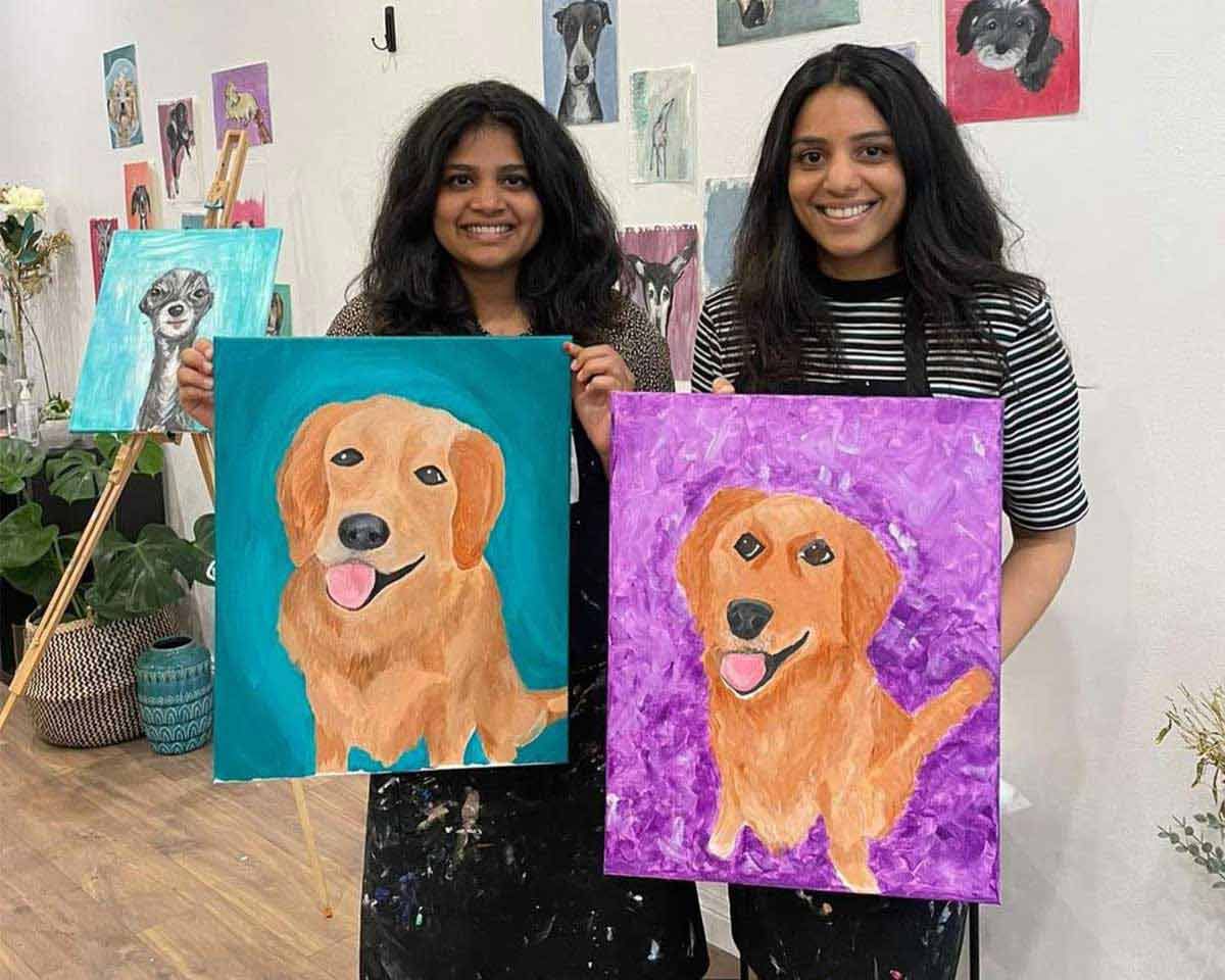 Two women holding up paintings of dogs in a Pinot and Picasso art studio session paint your pet.
