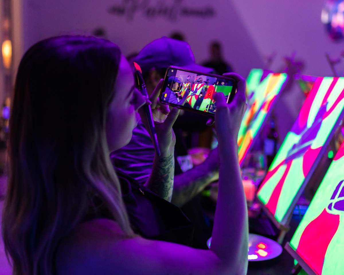 a person taking a photo on their phone of the neon nights artworks a group of people are painting at Pinot & Picasso