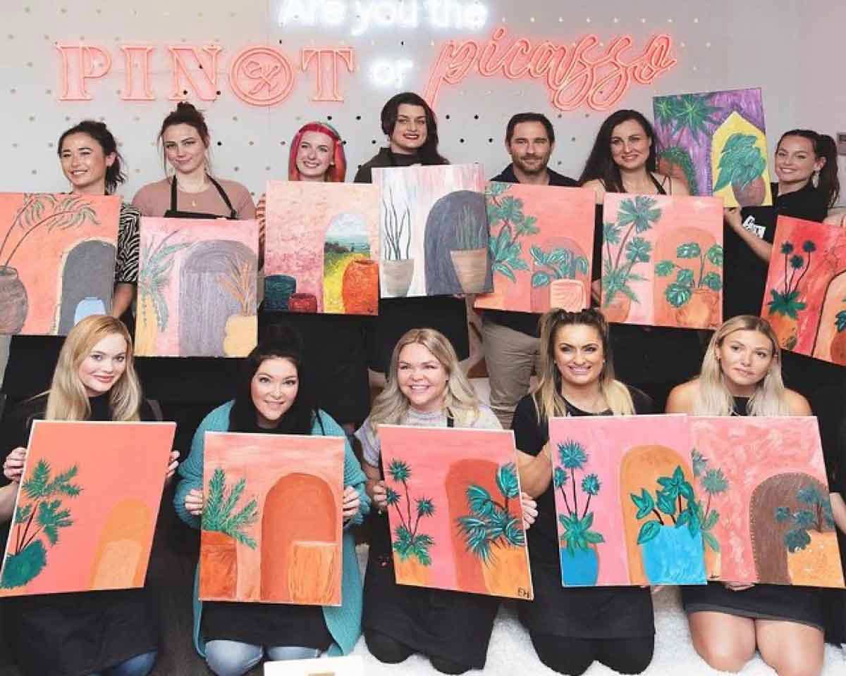 a group of people standing in front of a neon sign saying Pinot & Picasso with their plants over people artworks