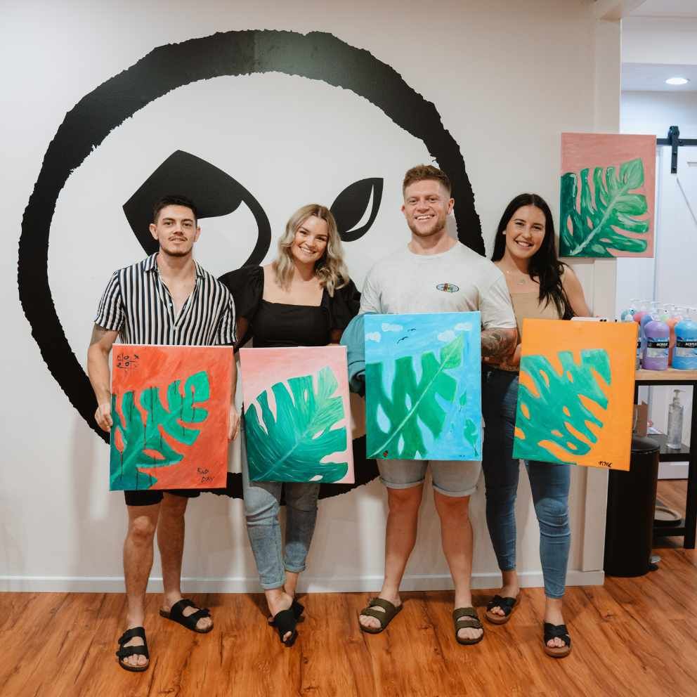 a group of people proudly holding up their artworks of the mosteria plant at a Pinot and Picasso studio