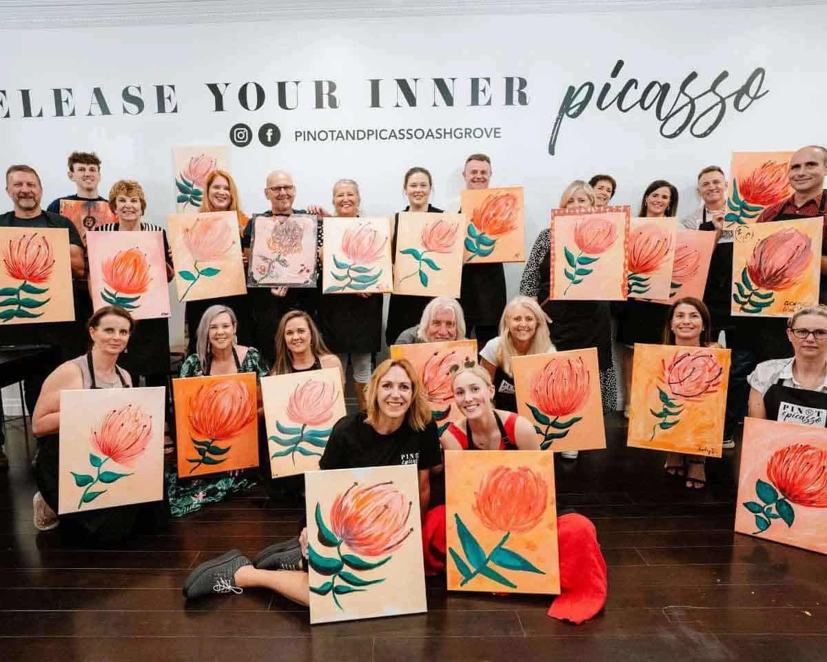 A group of people painting and posing with their artwork at a Pinot & Picasso paint and sip event.