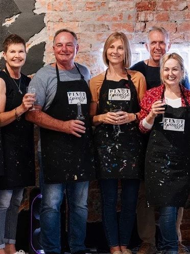 a group of people smiling with their aprons on and a glass of wine in their hands at a Pinot & Picasso session