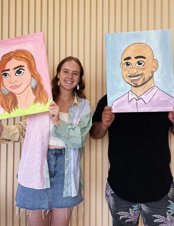 A couple participating in a Pinot & Picasso paint and sip event, holding up Disney animated painting style portraits of each other.
