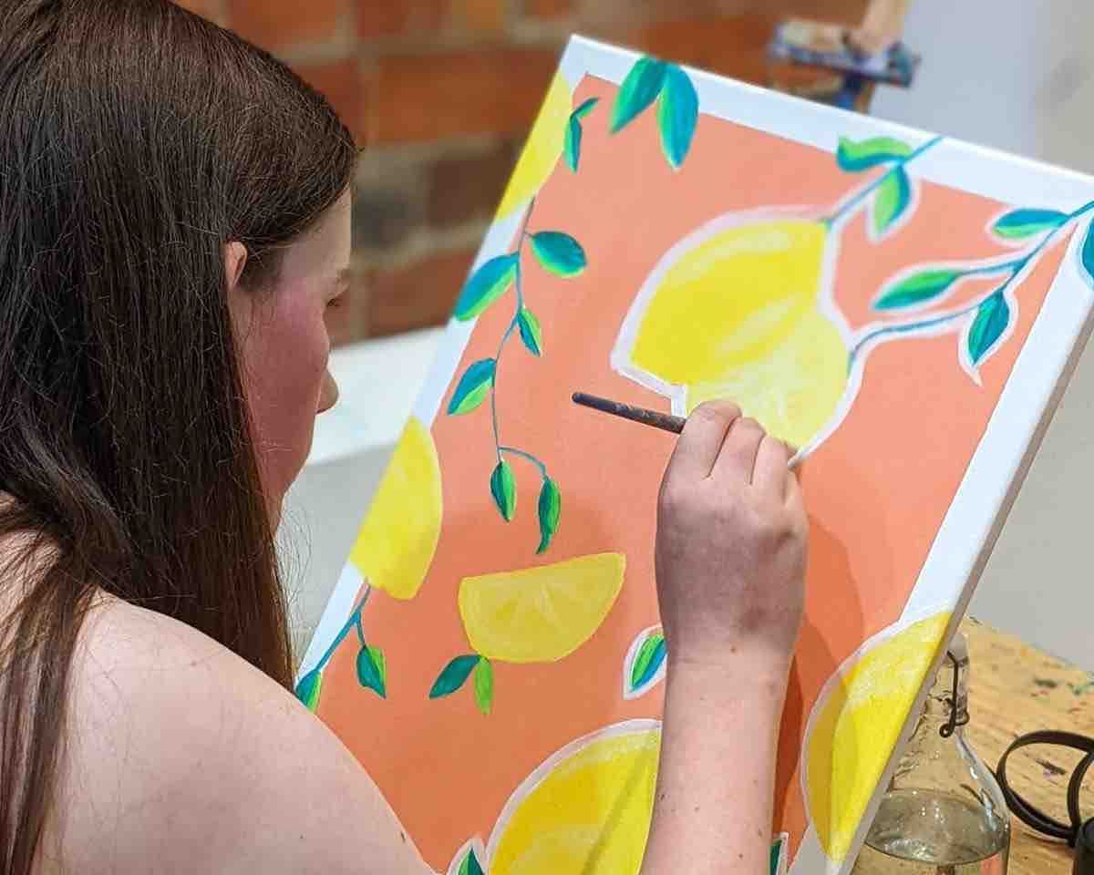 a woman painting lemons onto a canvas at a Pinot and Picasso session